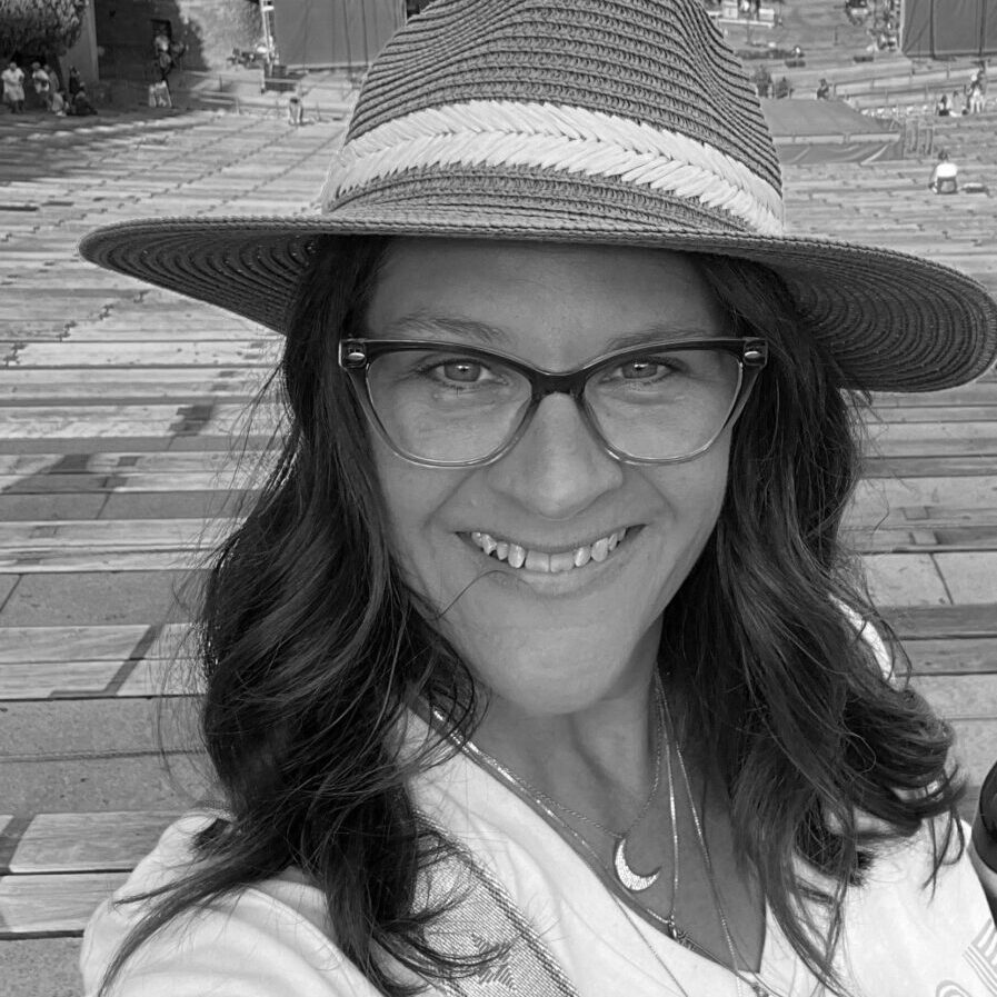 Smiling woman wearing glasses and a wide-brimmed hat outdoors.